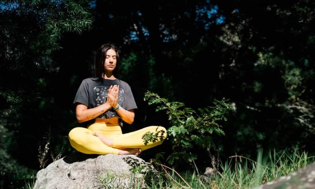 A woman is sitting on a large rock in a forest doing yoga