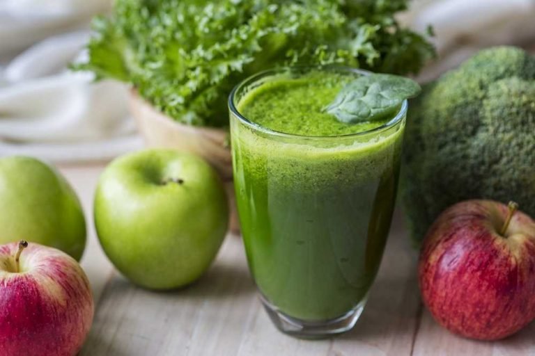 A Glass full with green juice and with some apples laying around