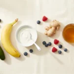 Overhead flat-lay of debloating foods including cucumber, banana, yogurt, ginger, berries, and herbal tea on a cream linen background